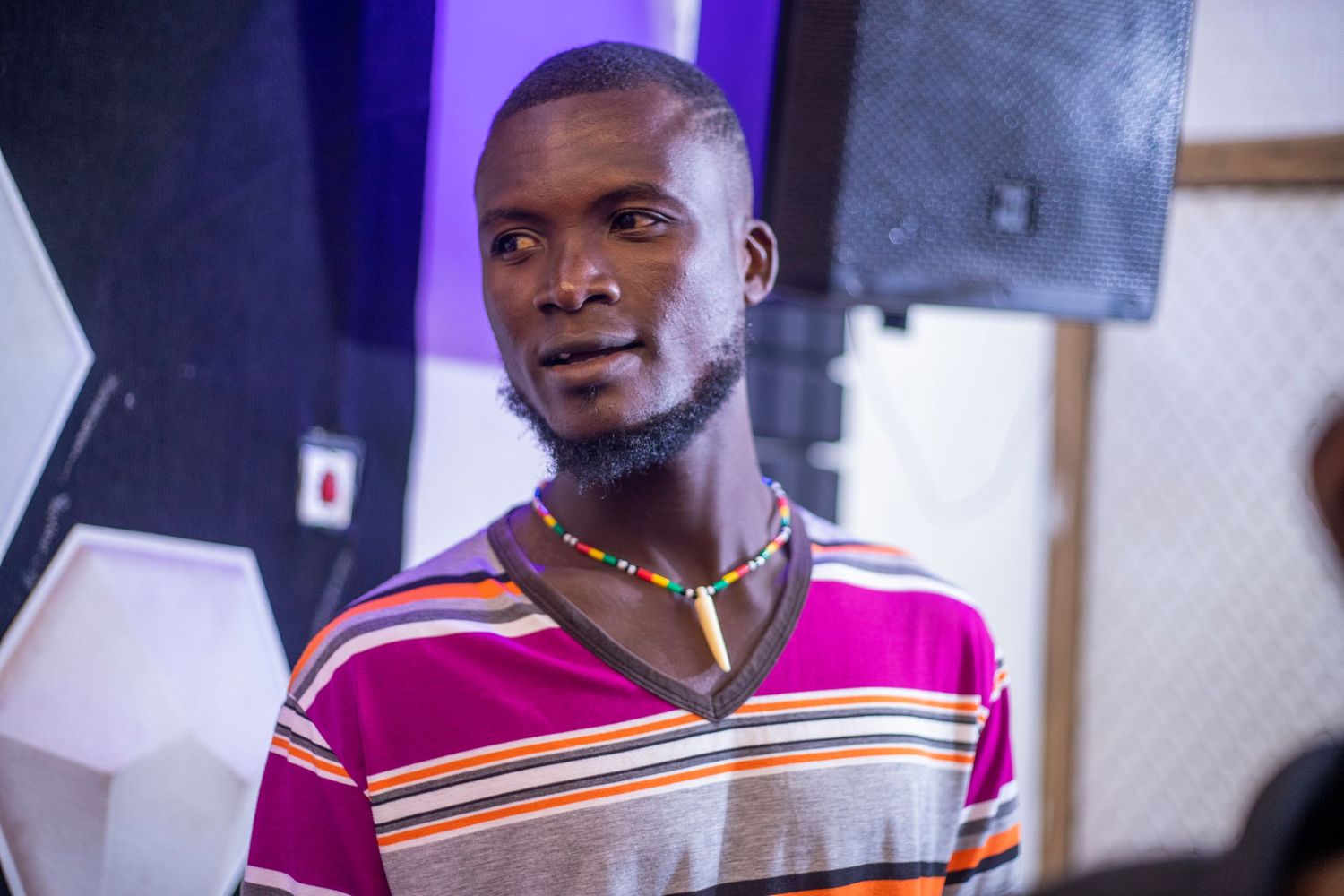 Young African man wearing colorful striped shirt and beaded necklace standing indoors in a studio setting.