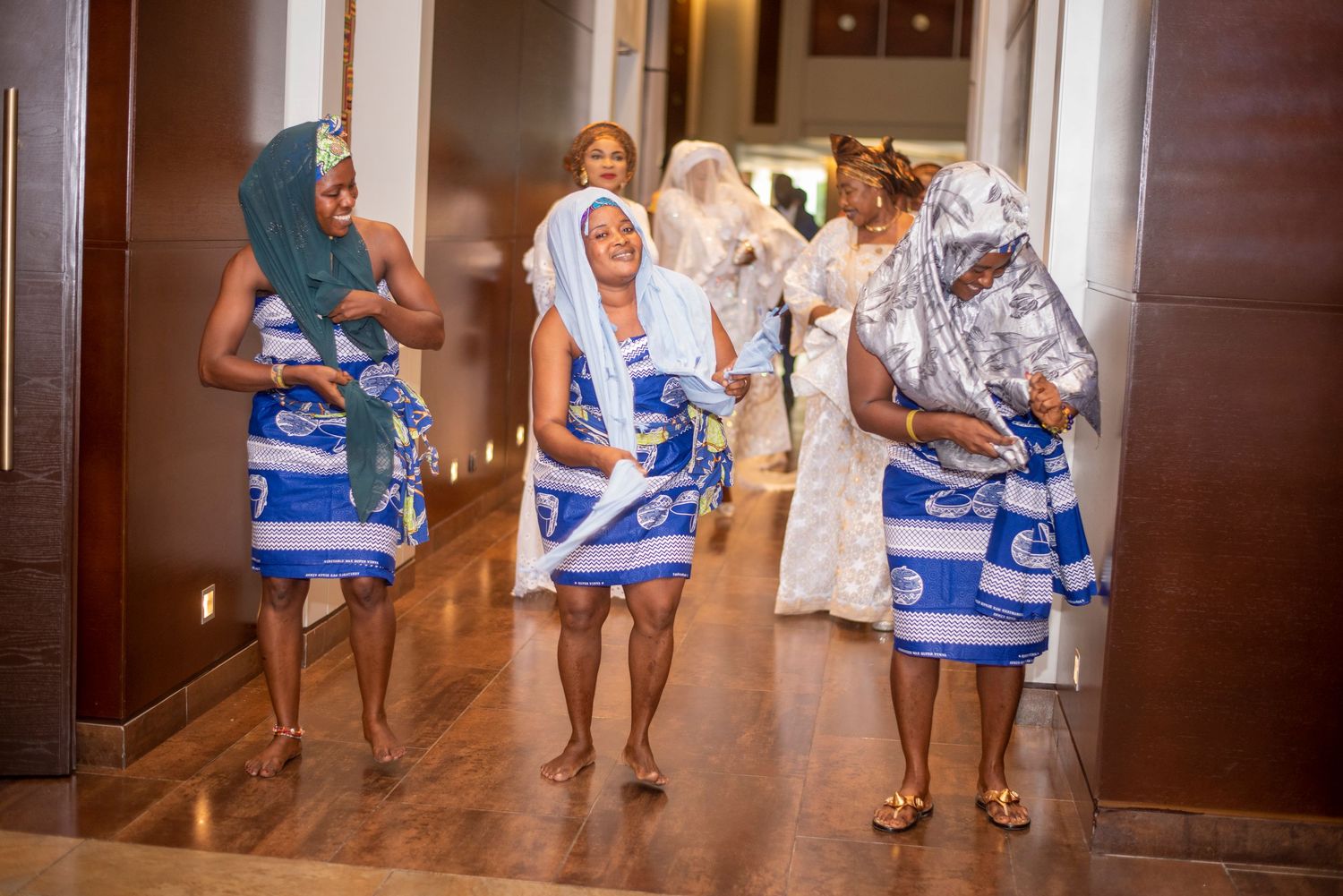 Group of African women in matching blue and white traditional attire walking through an elegant hotel corridor.