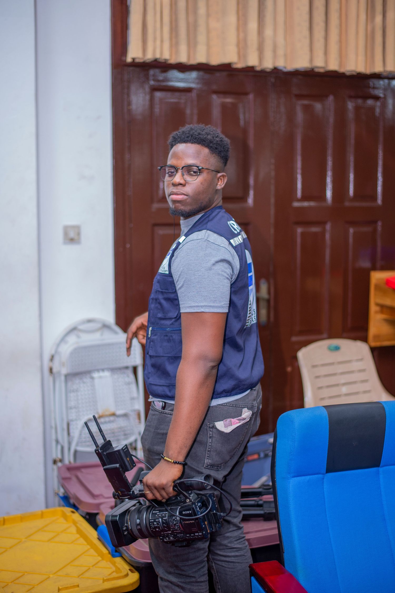 Young man with glasses holds a professional video camera while standing near colorful chairs indoors.