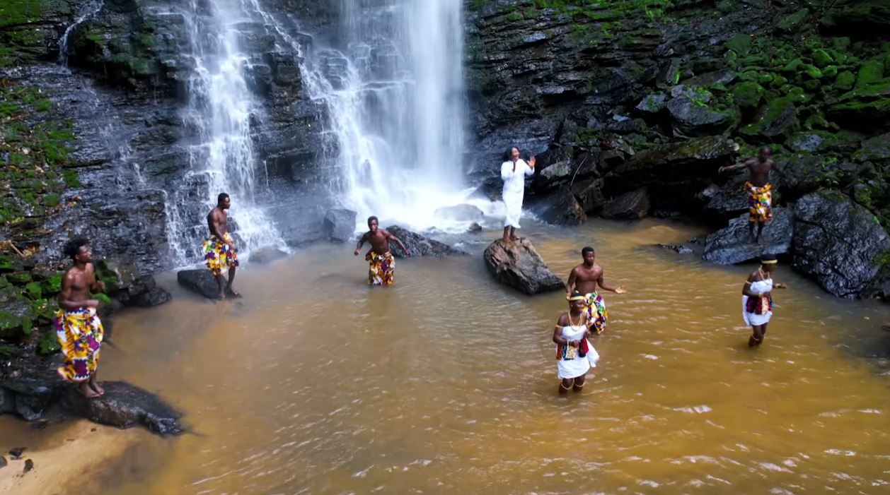 Group of traditionally dressed African dancers at base of scenic waterfall.
