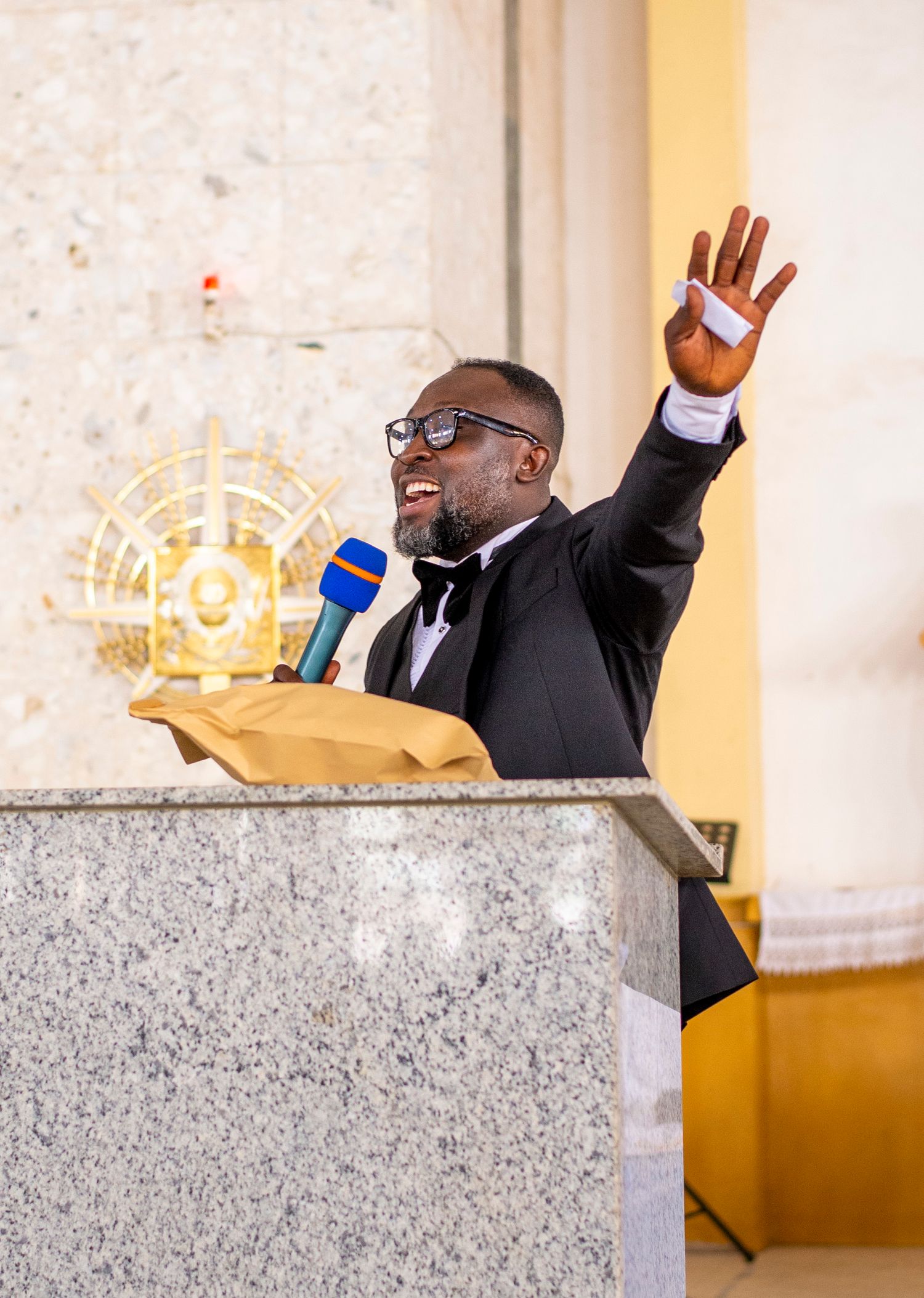 Man in black tuxedo speaking at podium with microphone, raising hand enthusiastically during speech.