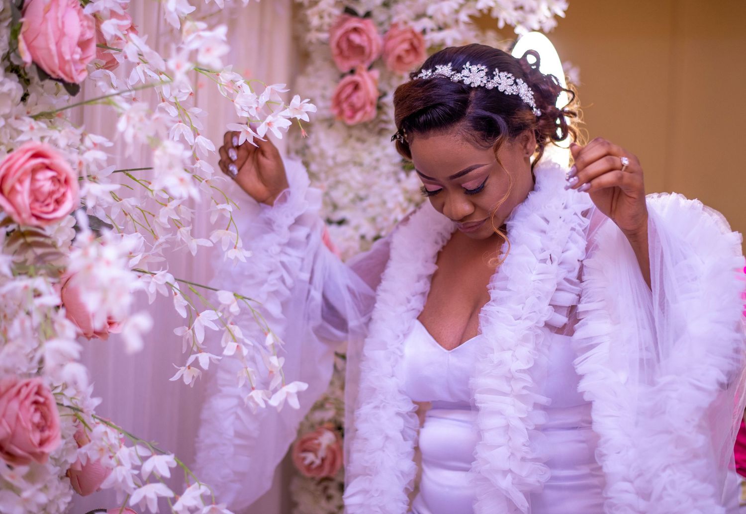 Elegant bride in white gown with fur stole adjusting hair accessory near pink floral backdrop.