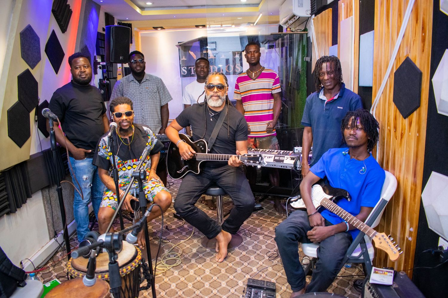 A group of musicians posing together in a recording studio with guitars and acoustic panels on the walls.