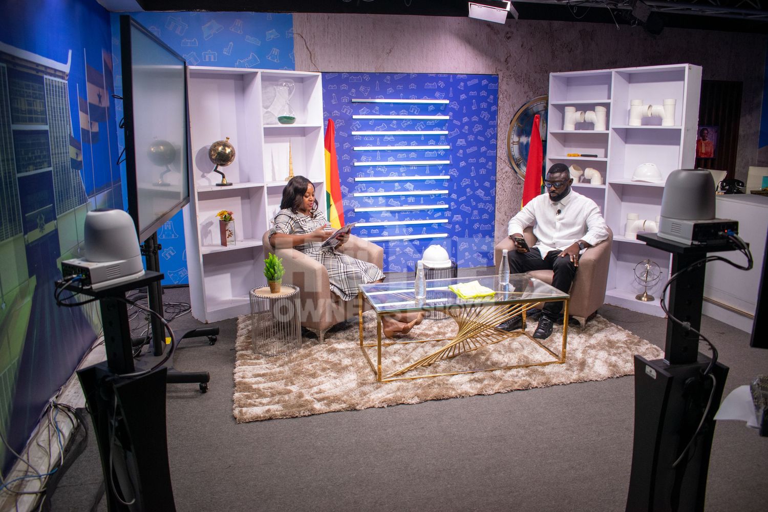 Two people sit across from each other on a TV talk show set with white bookshelves and colorful backdrop.