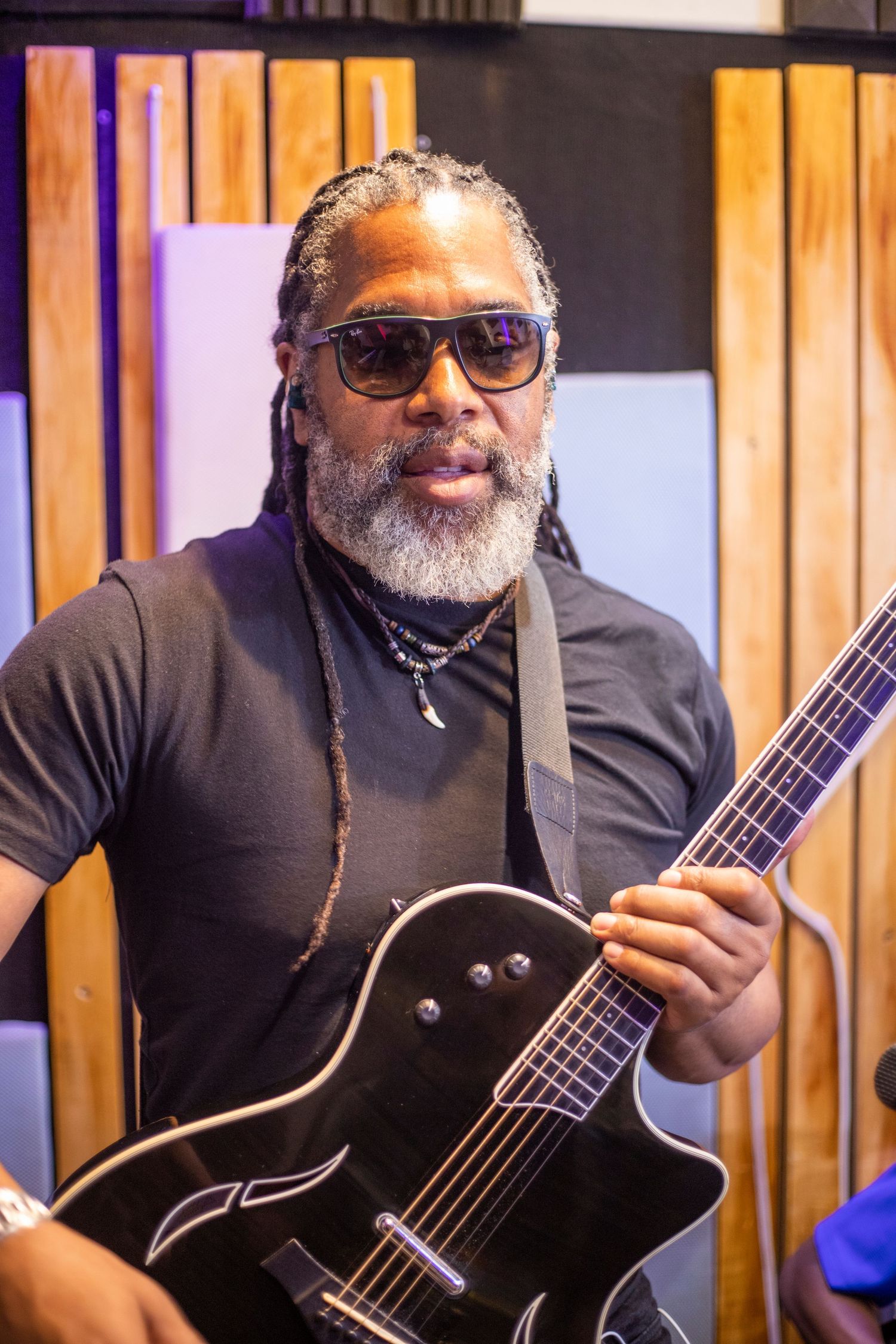 A musician with dreadlocks and sunglasses plays a black electric guitar in a recording studio.