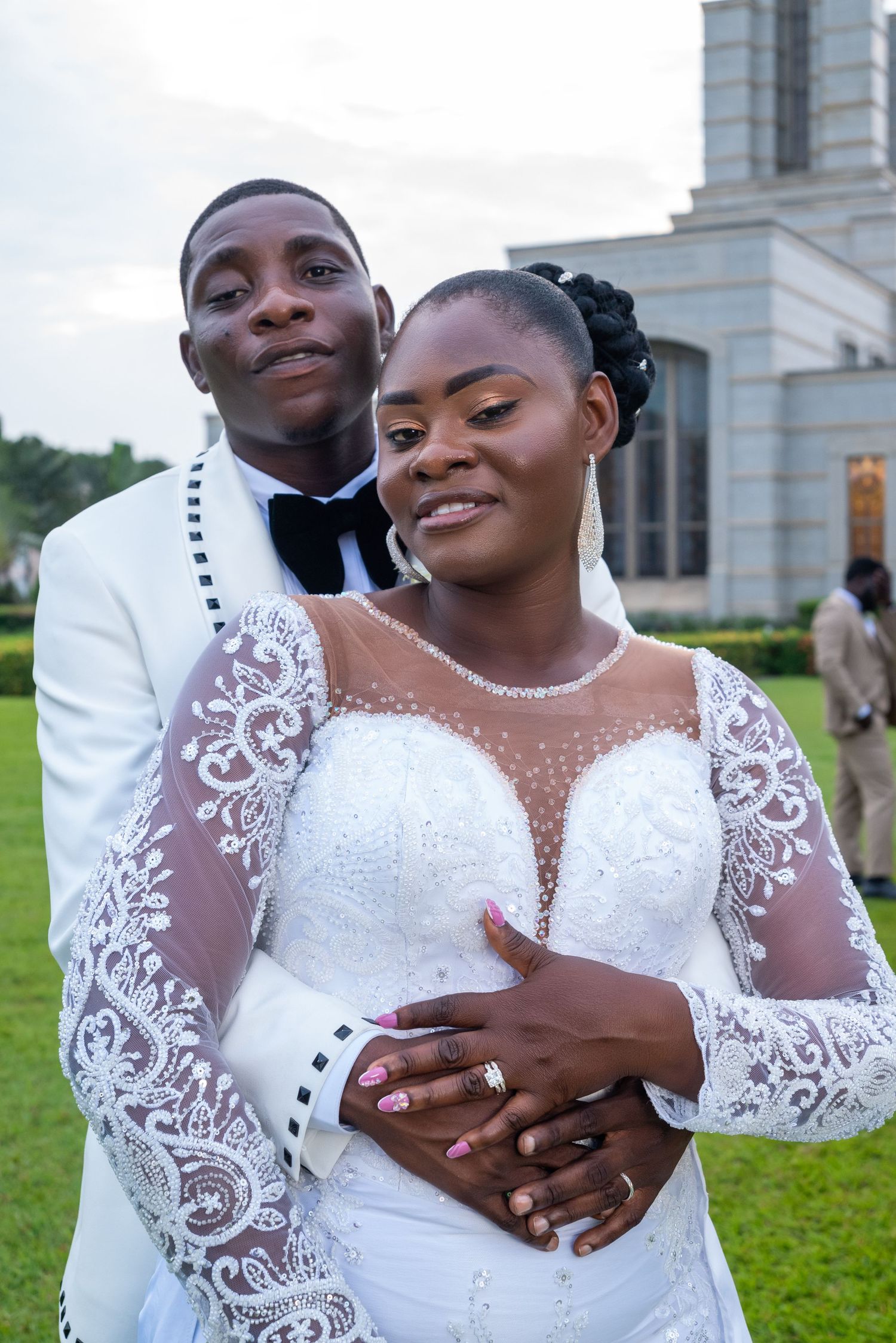 A couple in wedding attire poses outdoors, groom in white tuxedo embracing bride in lace gown.