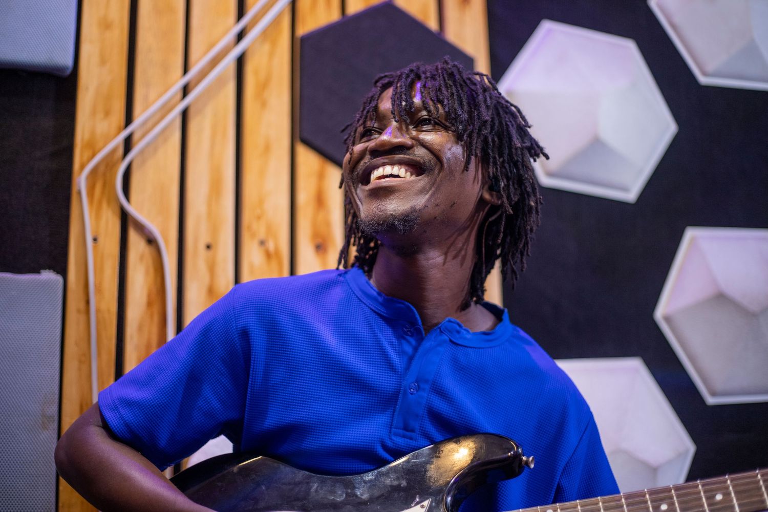 Smiling musician with dreadlocks playing electric guitar in blue shirt against geometric wall backdrop.