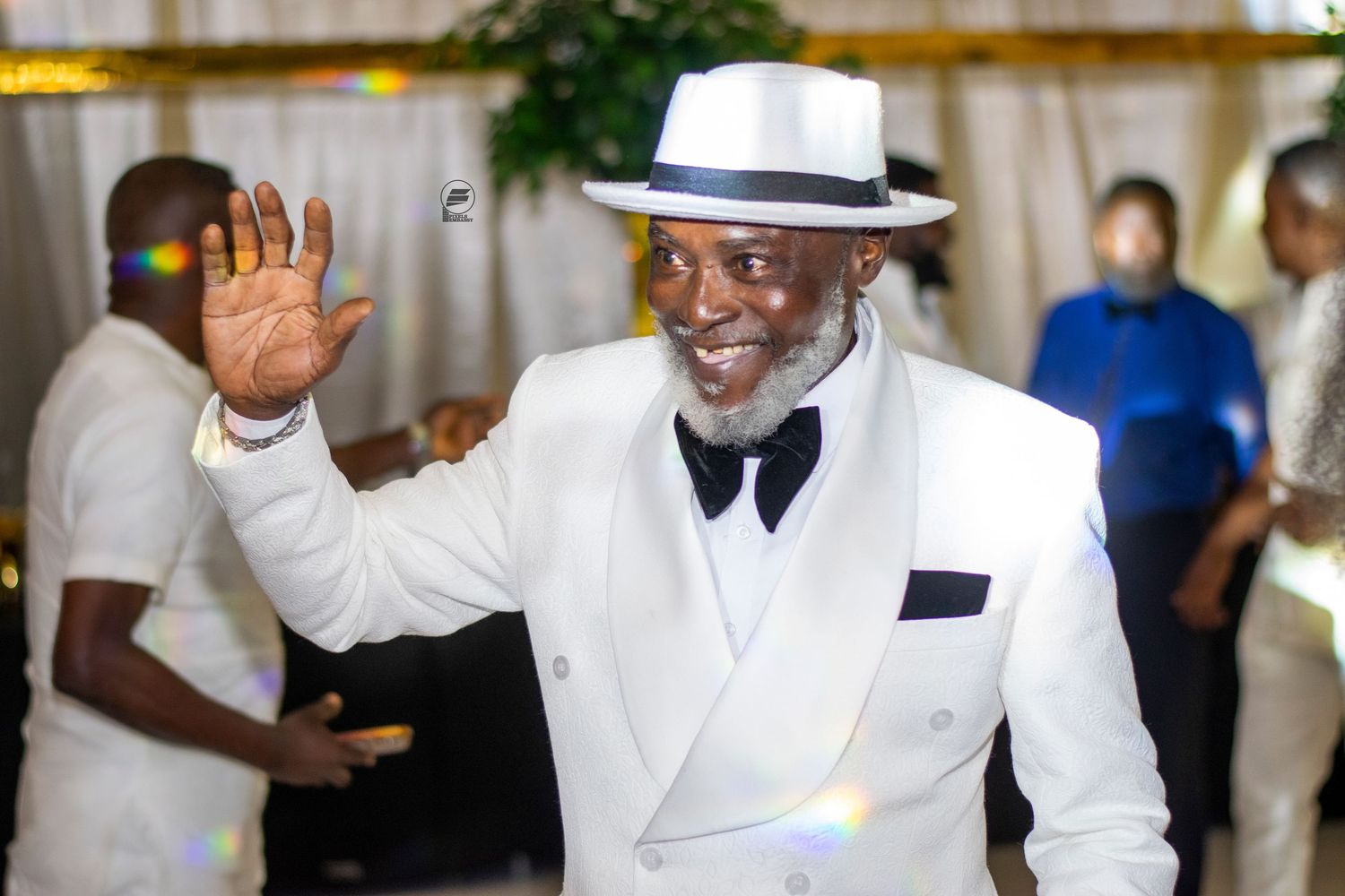 Elegant man in white suit and fedora hat waving at an indoor event, surrounded by guests in festive attire.