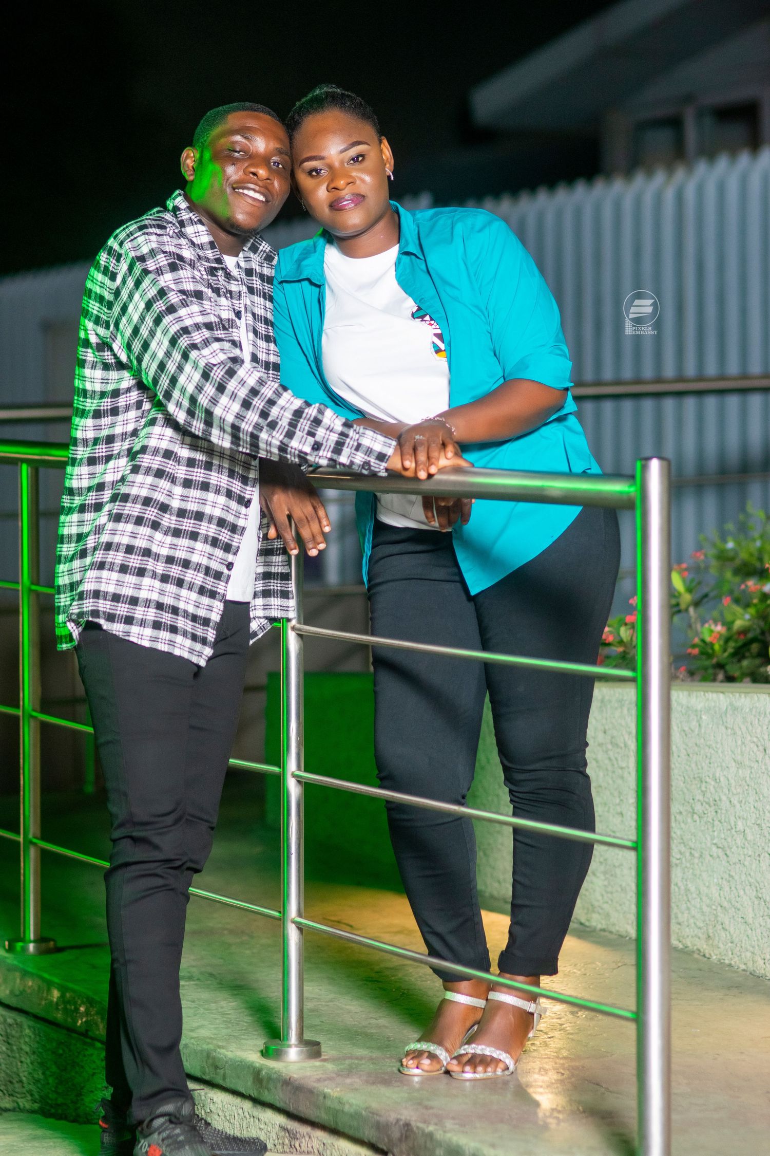 A smiling couple poses together leaning on a green railing outdoors at night, wearing casual attire.