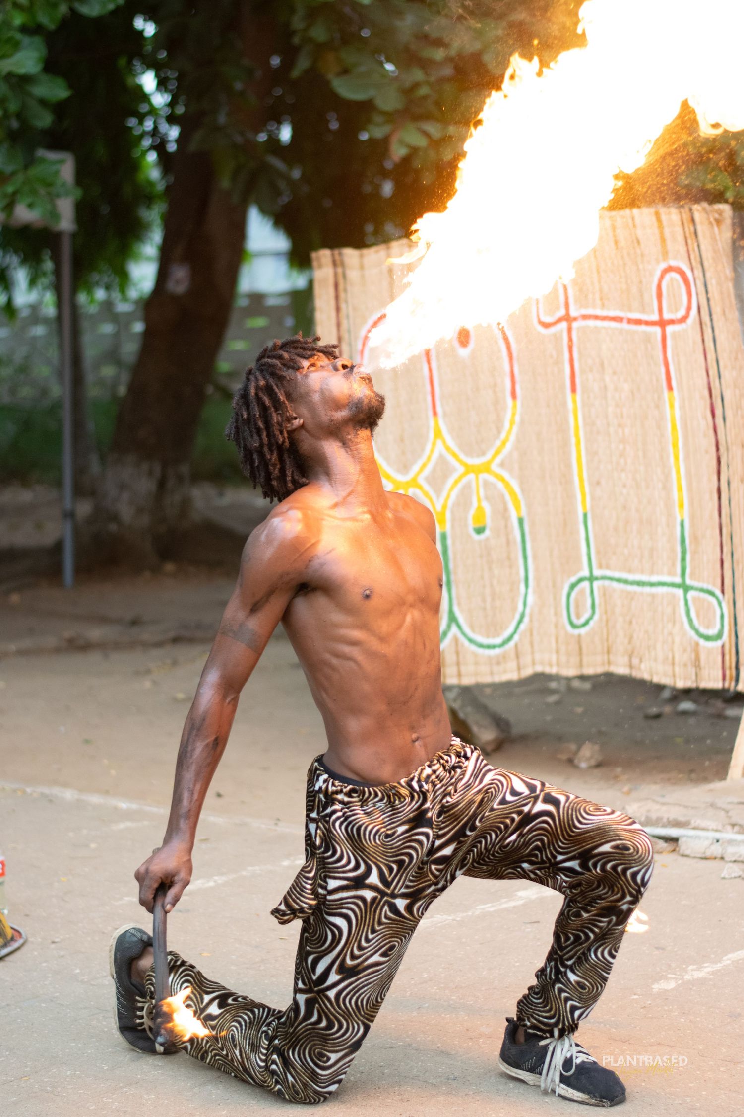 Athletic fire breather performing dramatic fire show outdoors against decorative background.