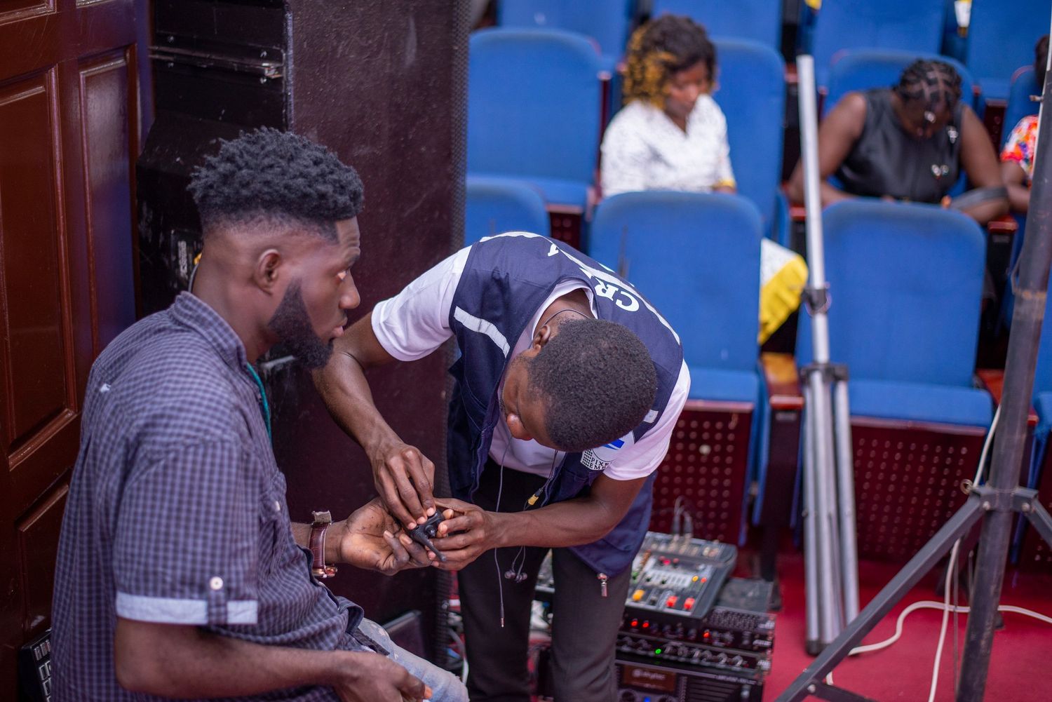 Two men working with audio equipment in a blue-seated auditorium during an event setup.