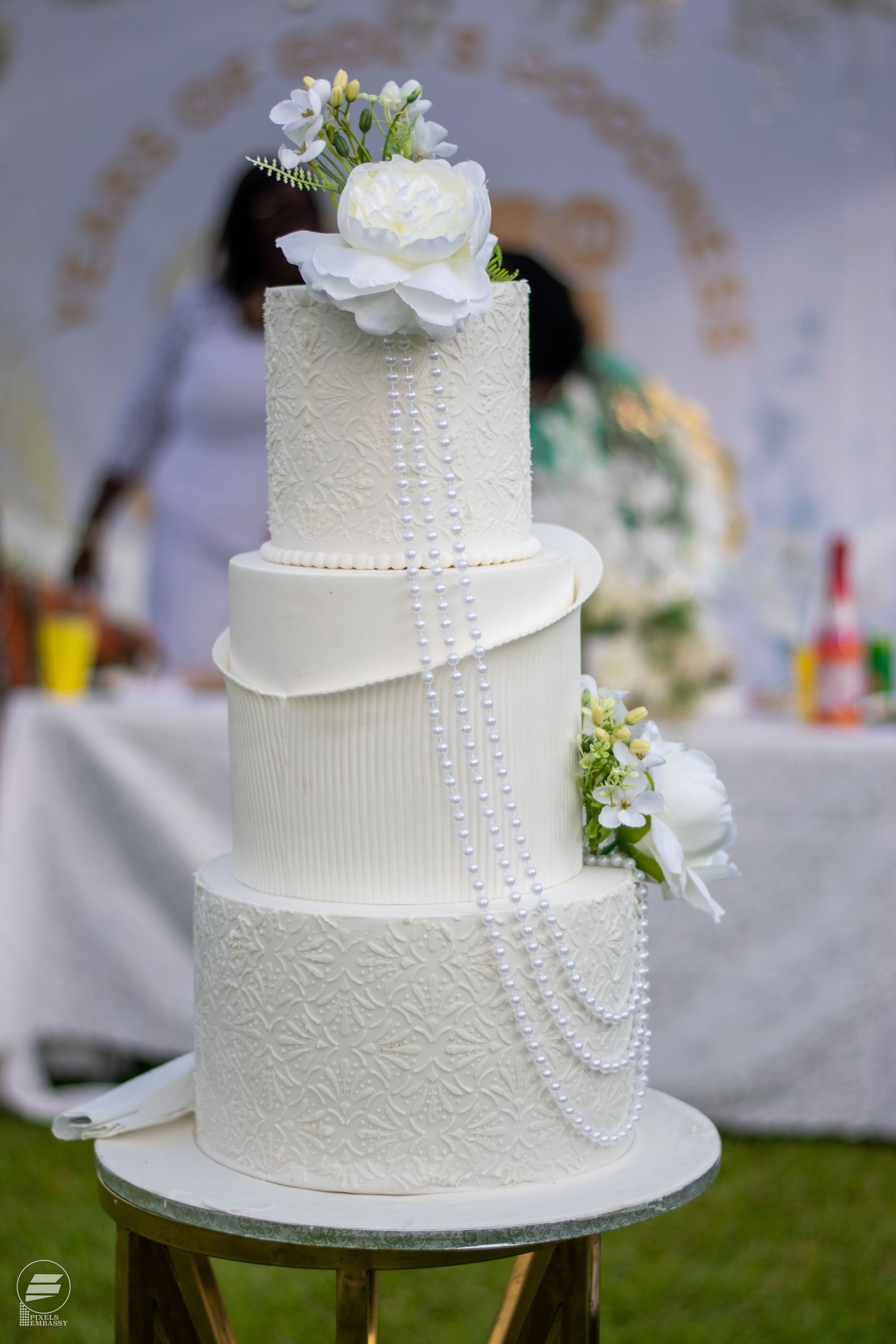 Elegant three-tier white wedding cake with pearl draping and white floral decorations outdoors.