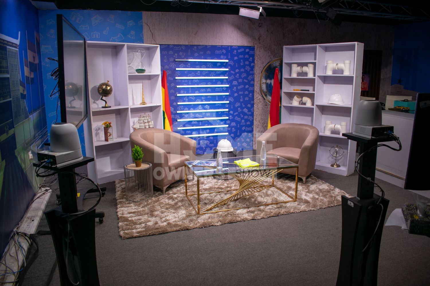 Empty TV talk show set with two beige chairs, gold coffee table, white bookshelves, and colorful backdrop.