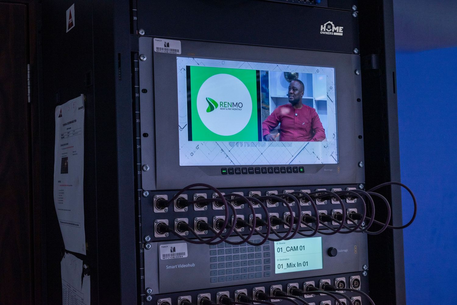 A rack-mounted AV system displays a video feed of a person on a small monitor with a green logo screen.
