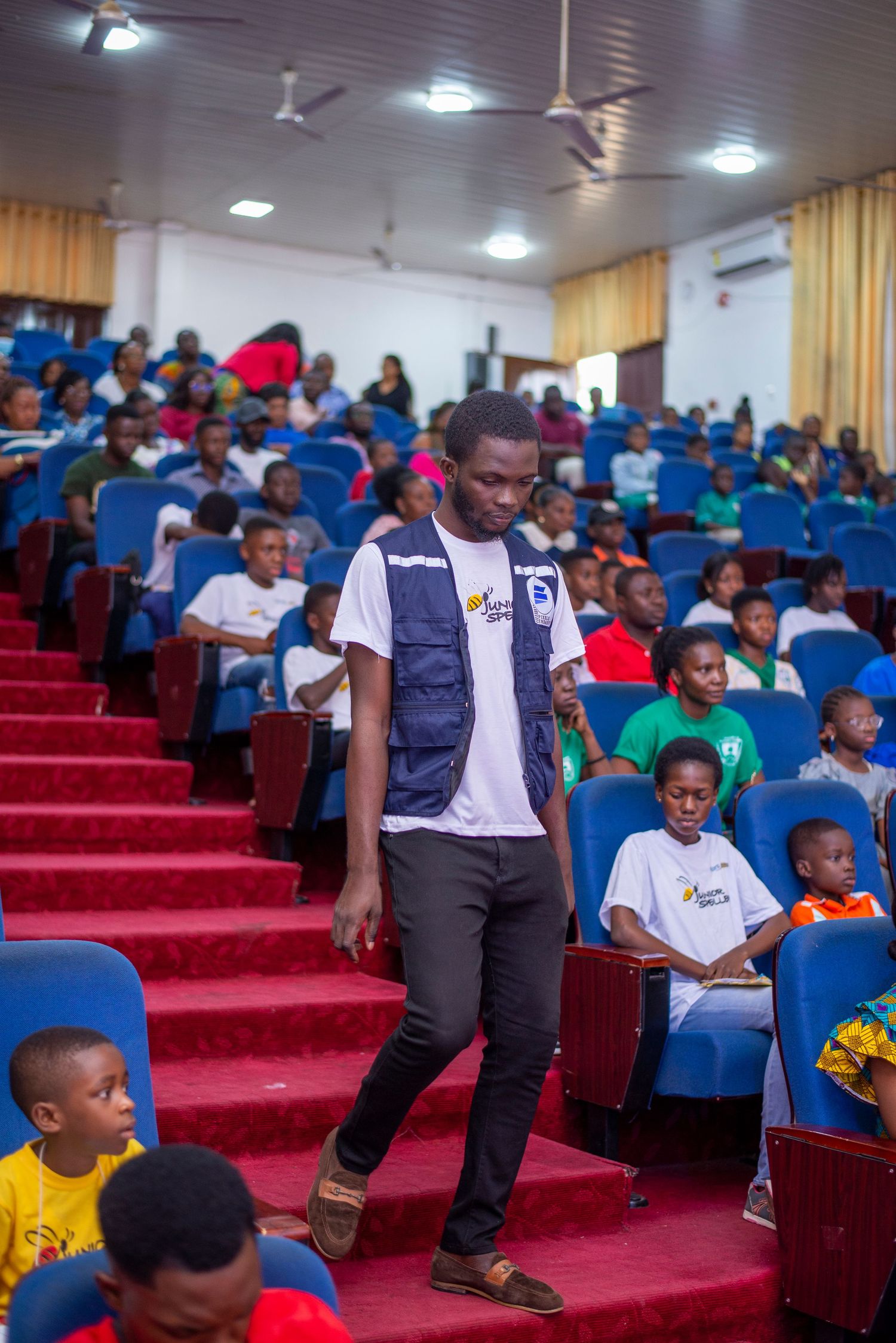 Man in blue vest walking through a crowded blue-seated auditorium during an event.