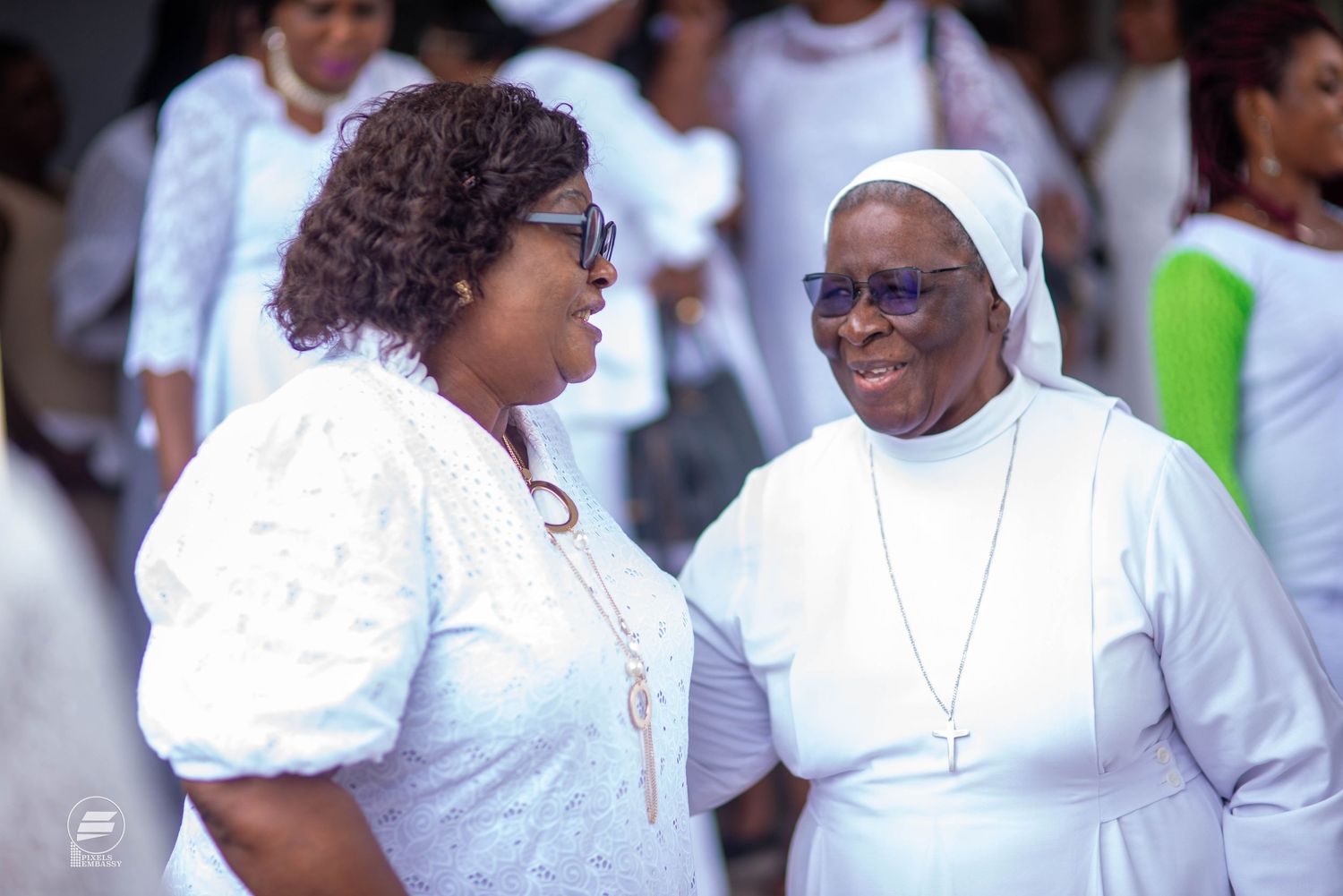 Two African women in white attire conversing outdoors, one wearing a nun habit with a cross.