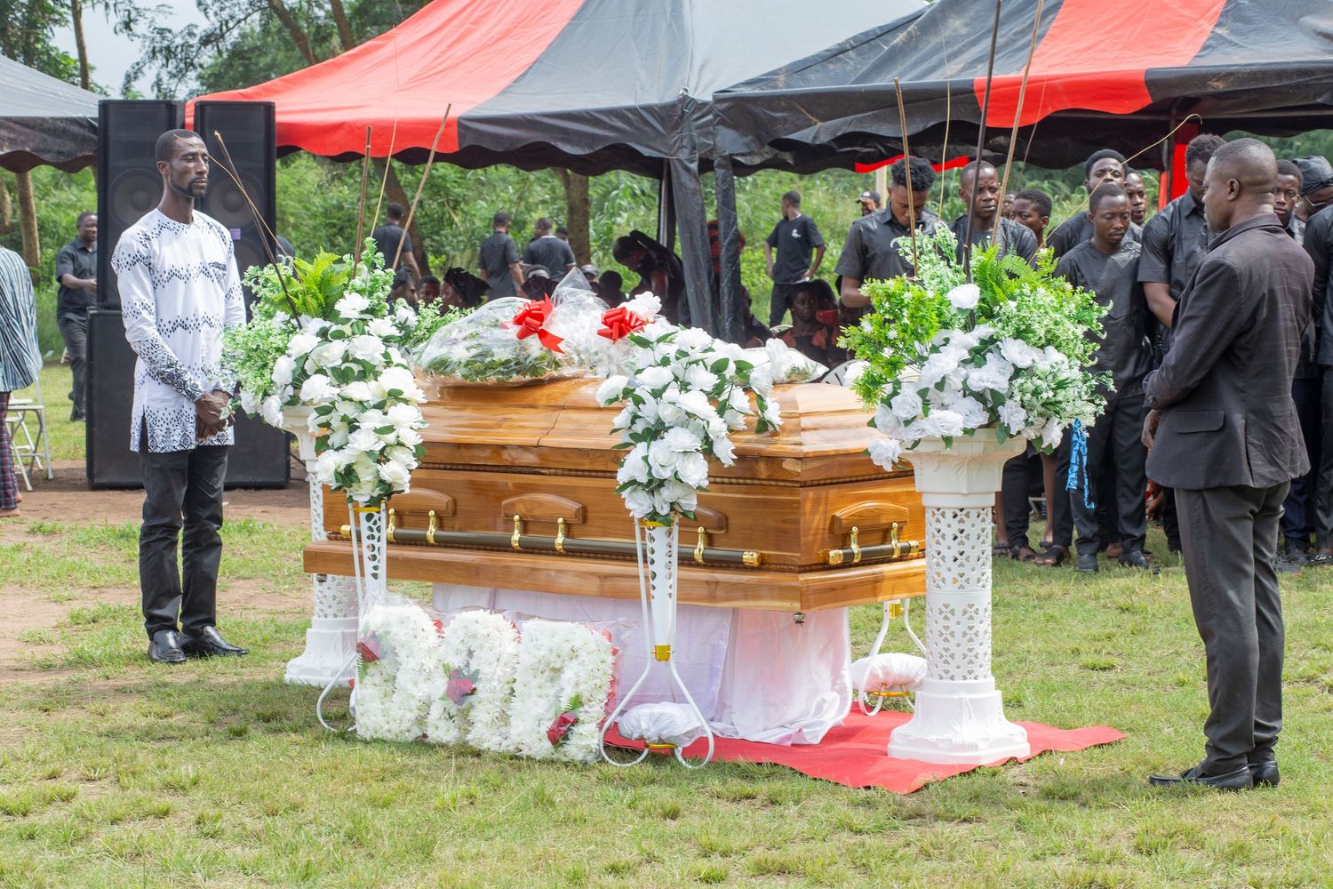 Funeral ceremony with two wooden caskets surrounded by white floral arrangements under a red tent.