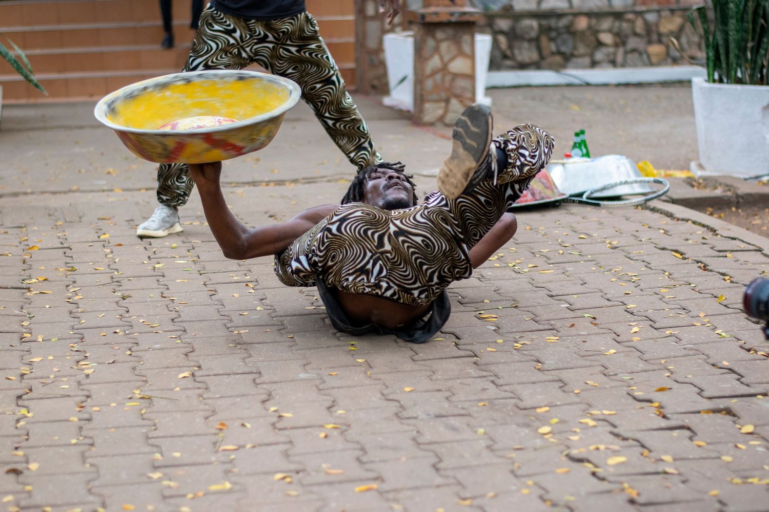African street performer in camouflage doing an acrobatic floor move while balancing a large bowl.