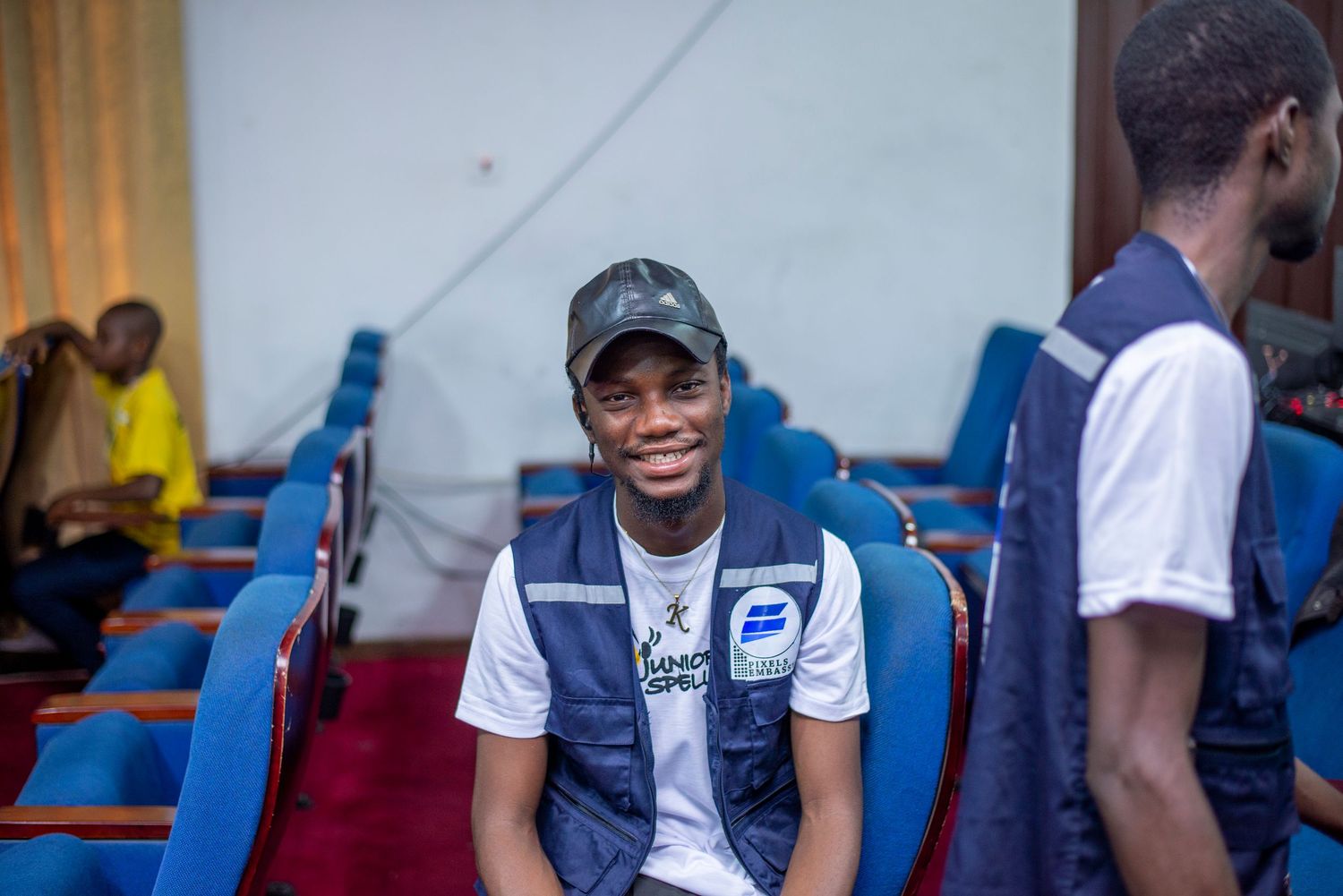 Smiling young man wearing a blue vest and cap sits in a blue auditorium chair at a Junior Speller event.