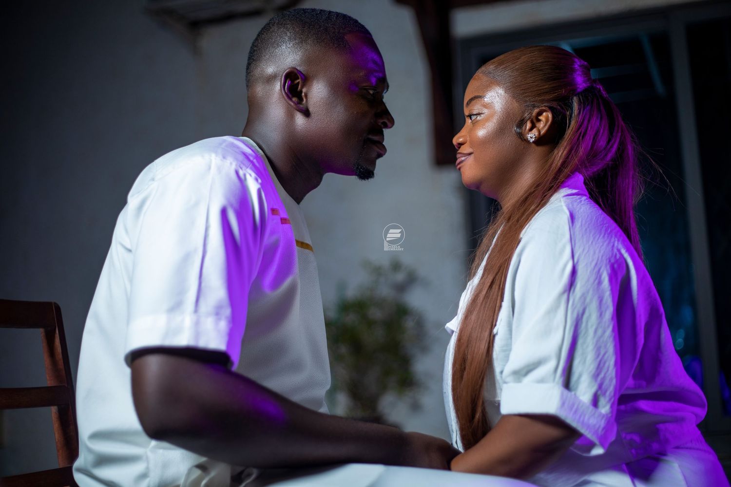 Young couple in white outfits facing each other in a romantic scene with purple lighting.
