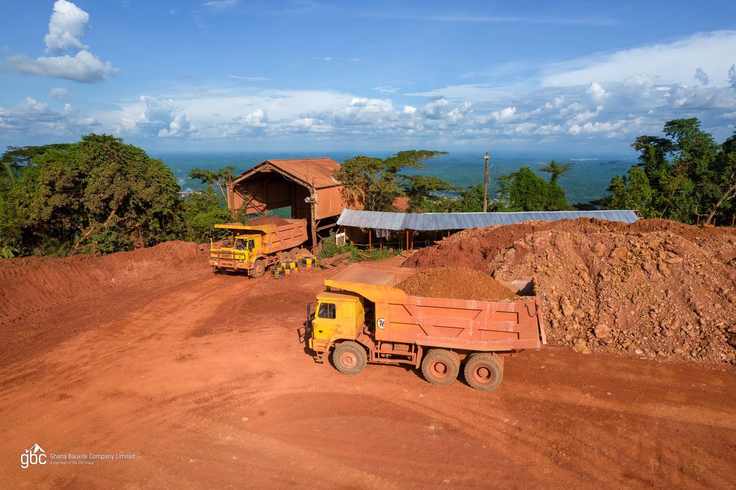 Yellow dump trucks on red soil construction site with aerial forest view.