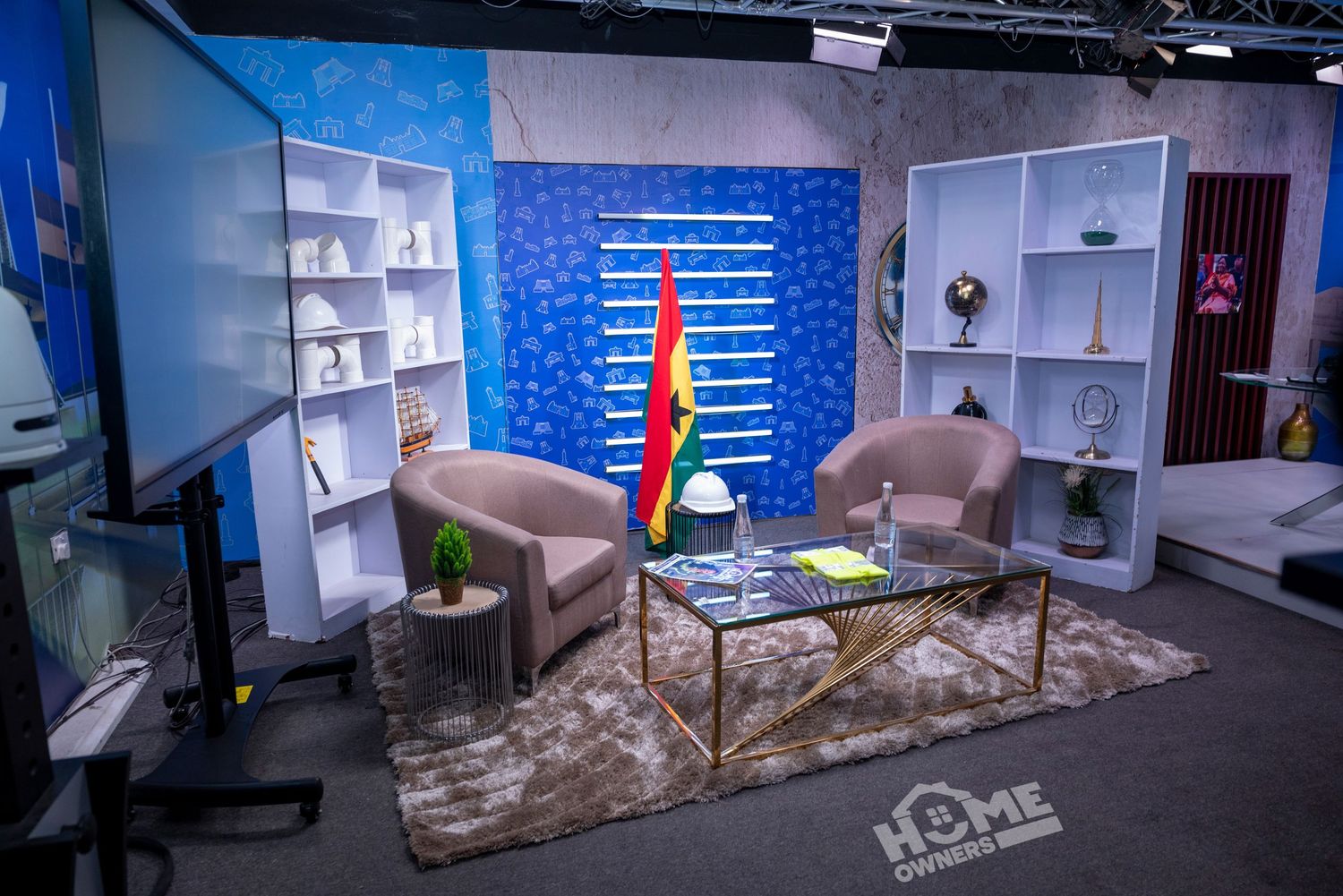 TV studio set with two beige chairs, glass coffee table, white bookshelves, and Ghana flag on blue backdrop.