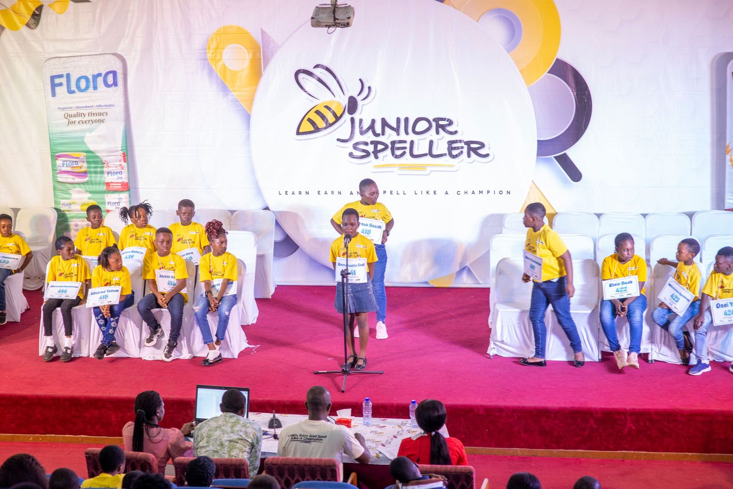 Child stands at a microphone on stage during a Junior Speller spelling bee competition event.