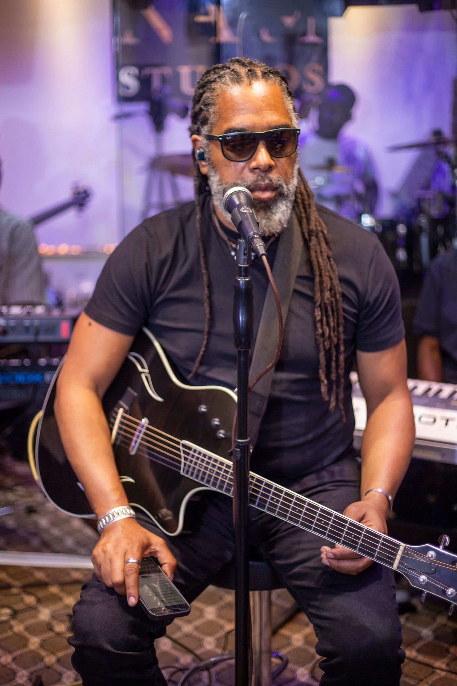 A musician with dreadlocks and sunglasses plays a black acoustic guitar at a microphone stand in a dimly lit studio.