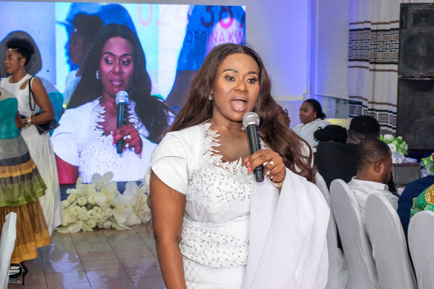 Elegant woman in white dress speaking into microphone at a formal event with projection screen behind.