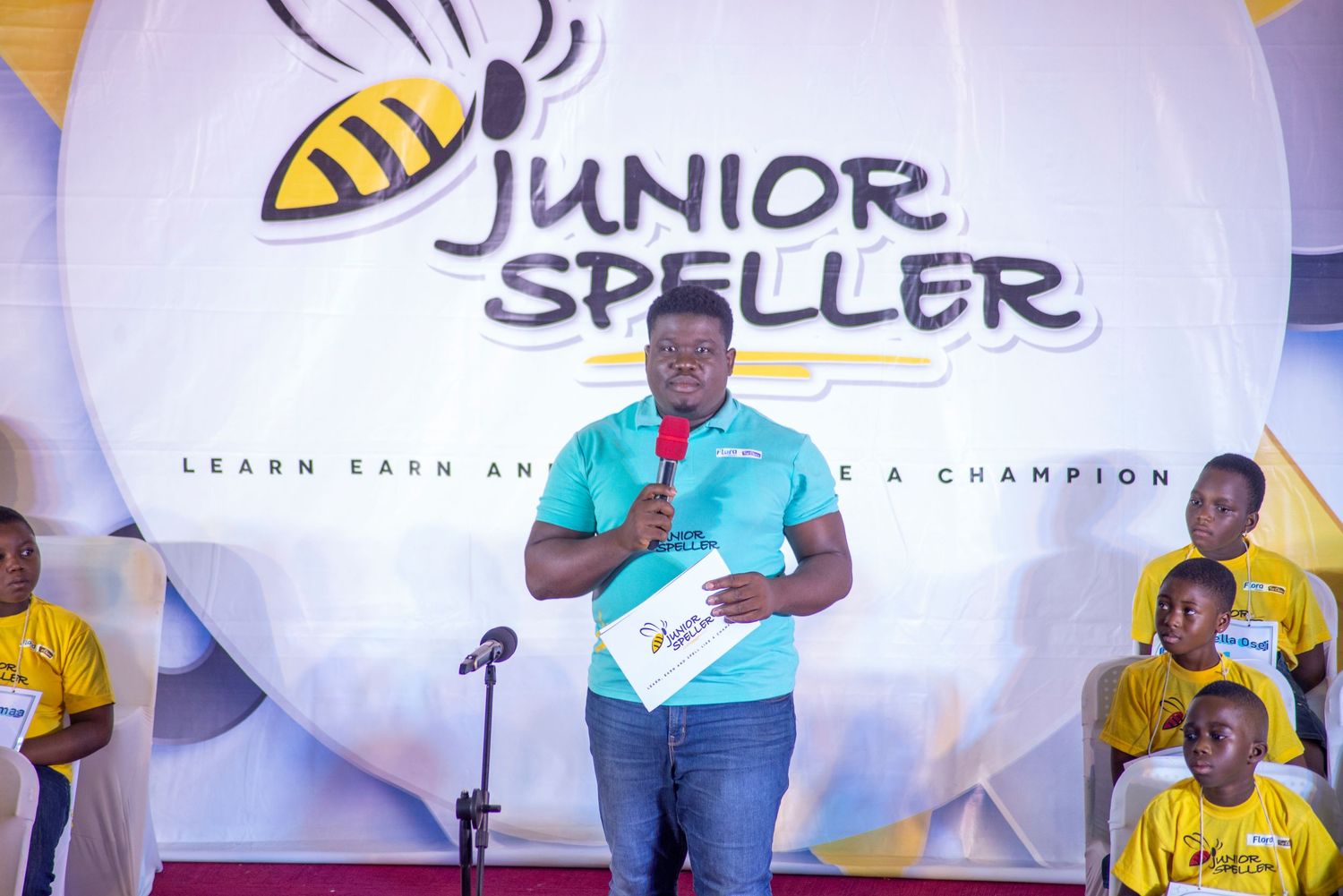 Man in a blue shirt speaks at a podium microphone at a Junior Speller championship event.