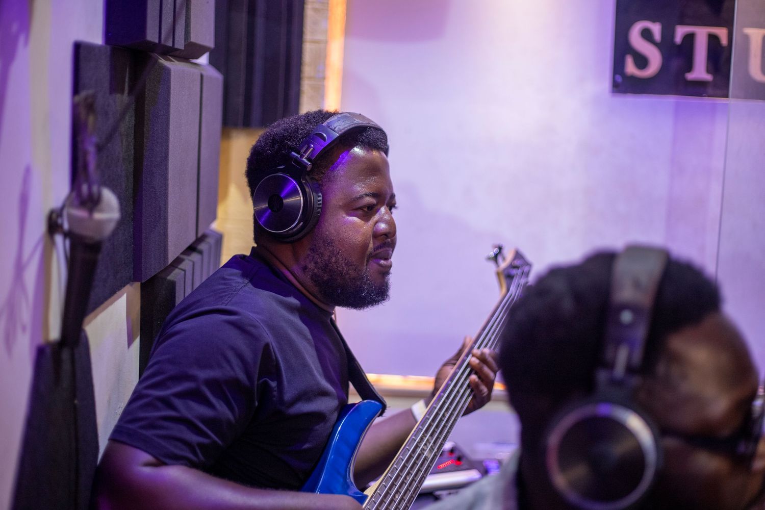 Musician wearing headphones plays bass guitar in a professional recording studio under purple lighting.