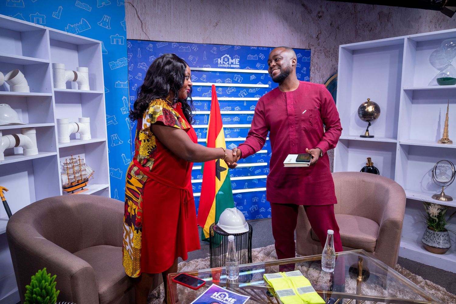 Two professionals in colorful attire shake hands in a modern office setting with shelves and flags in the background.