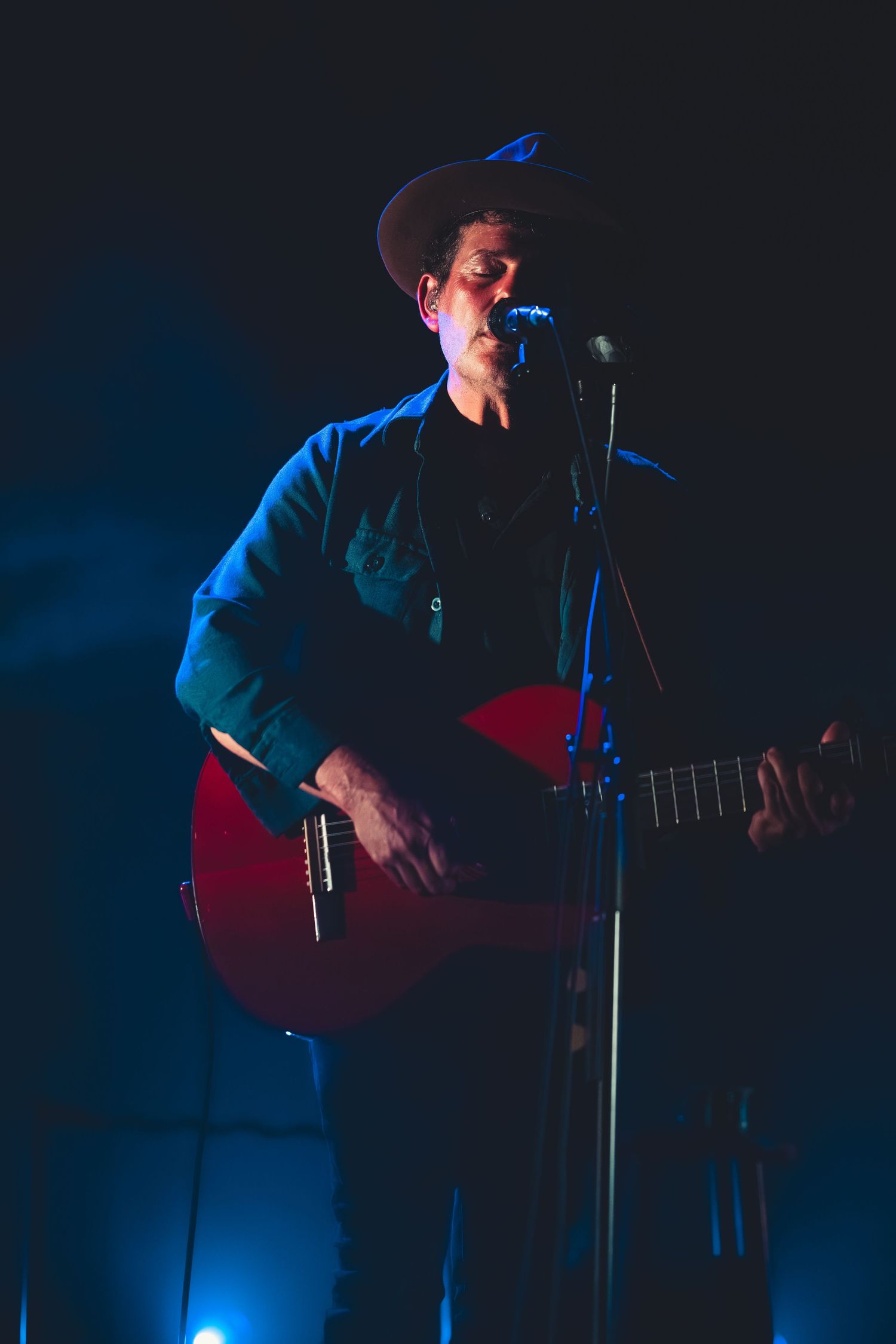 Male musician in cowboy hat singing into microphone while playing red acoustic guitar under blue stage lights.