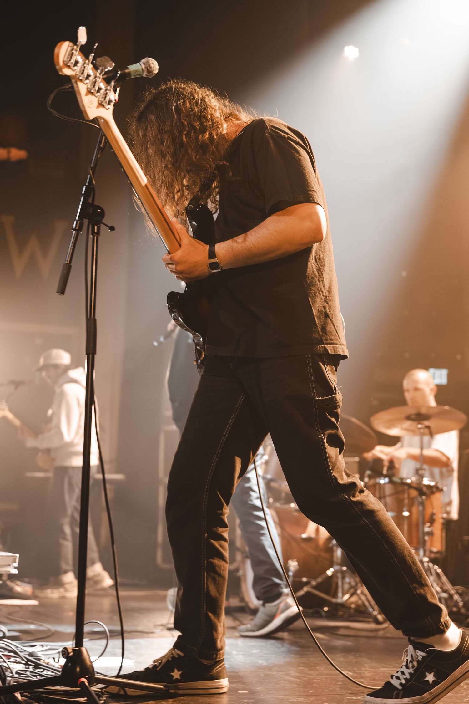Long-haired guitarist in black outfit performs on stage, leaning over electric guitar under warm amber stage lights.