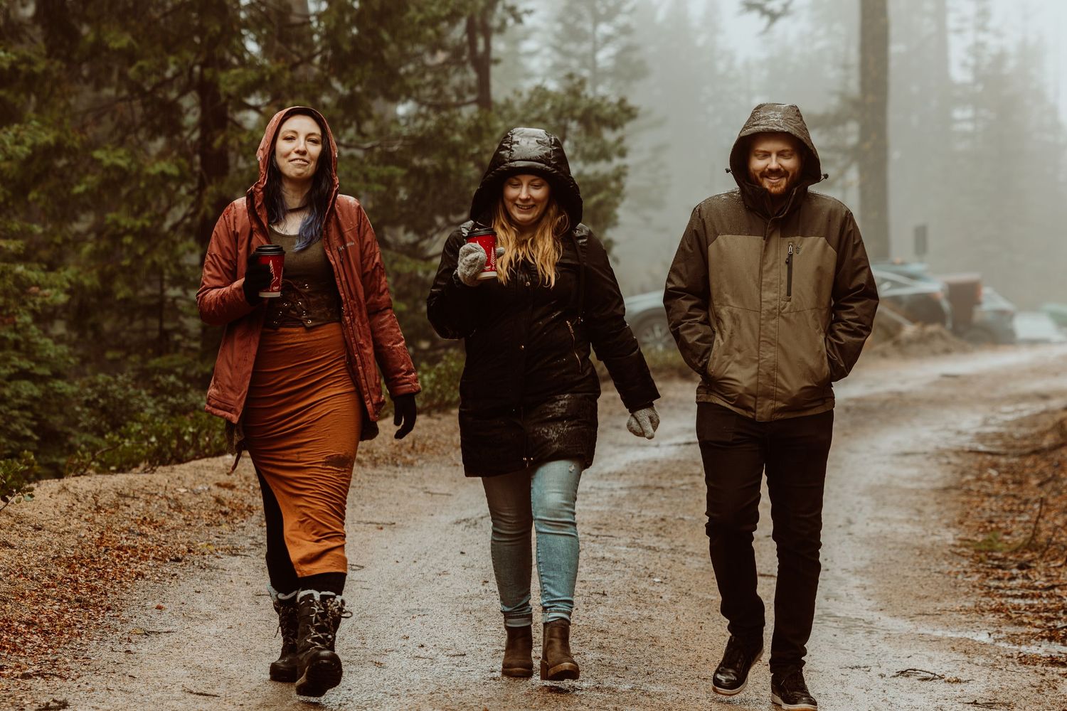 Three smiling friends walking together on a rainy forest path in outdoor gear.