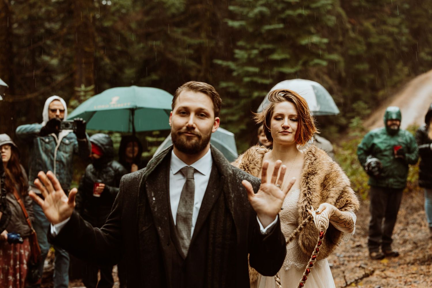 Groom raises hands while bride in fur stole stands beside him at rainy outdoor forest wedding.