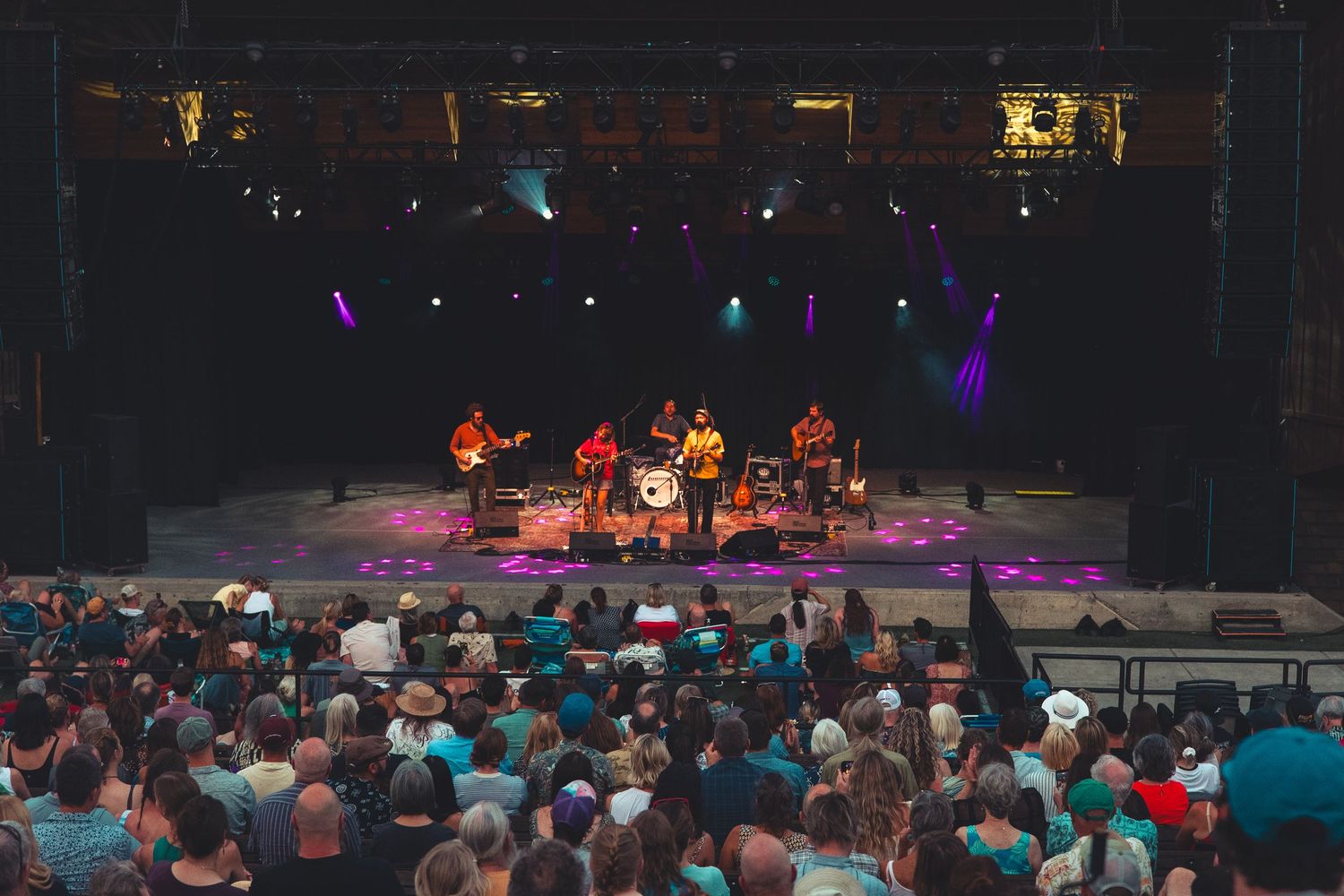 Live band performing on a colorfully lit stage with purple and gold lights for a large outdoor crowd.