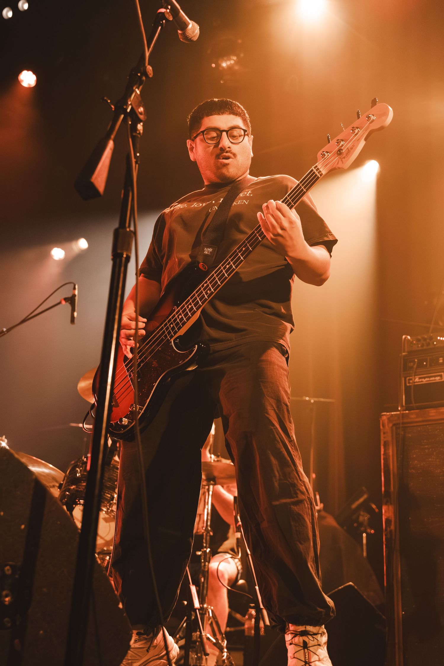 Male bassist with glasses plays bass guitar on stage under dramatic orange and white spotlights.