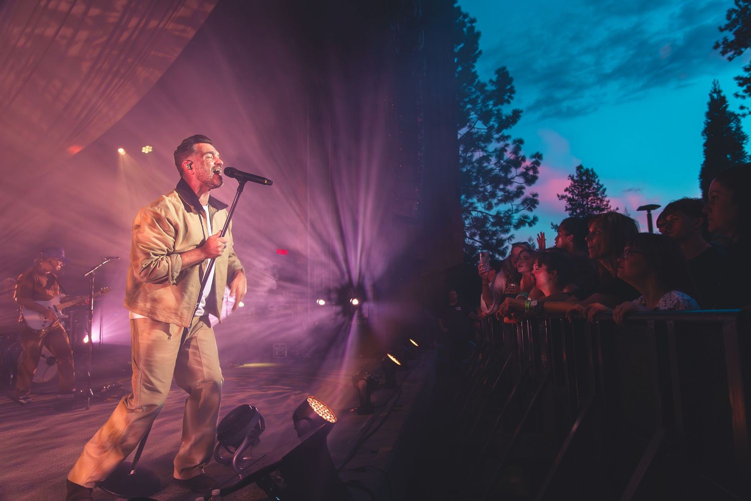 A male artist performs on an outdoor stage at dusk with a crowd gathered at the barrier.