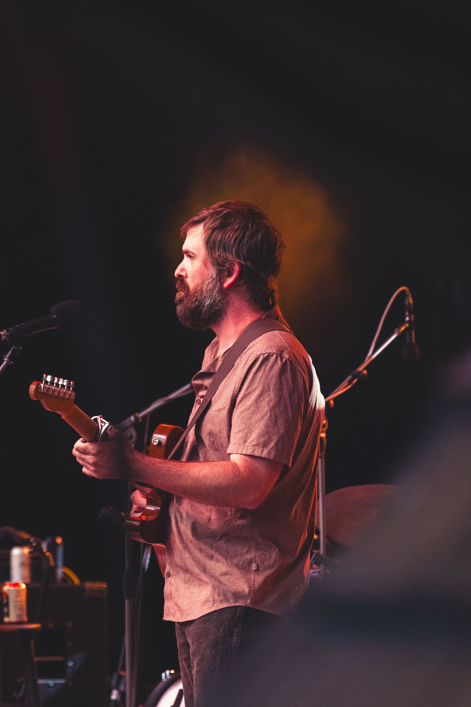 Bearded man playing acoustic guitar on stage under warm amber and yellow stage lighting.