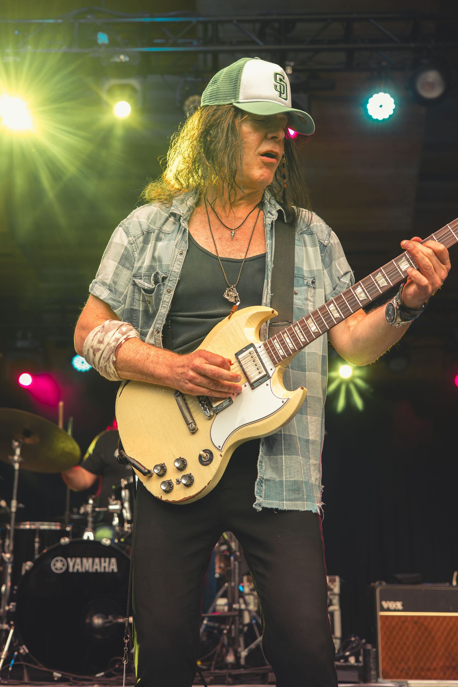 Long-haired guitarist in plaid shirt and cap playing white Gibson SG guitar on stage.