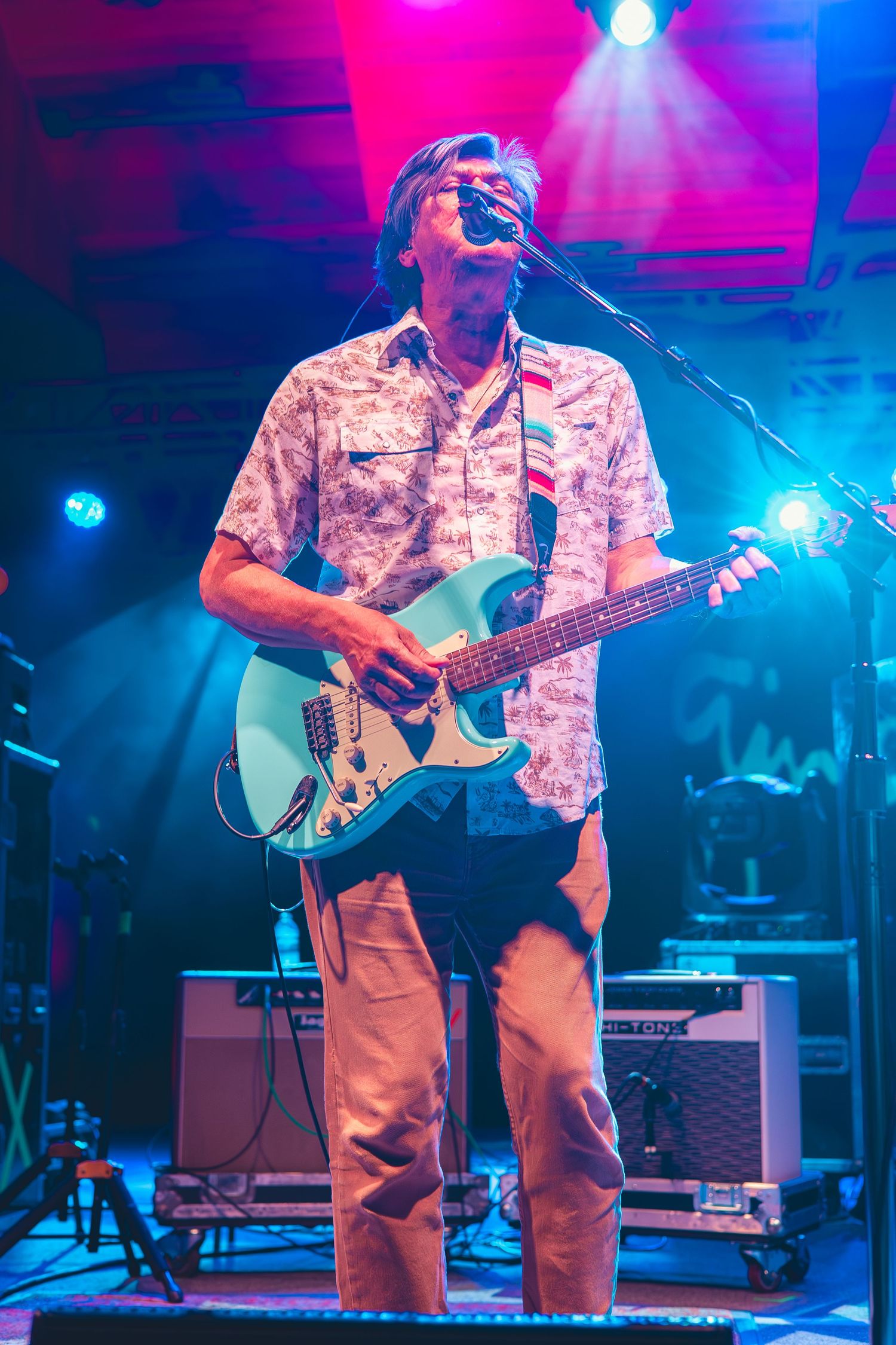 Guitarist singing into microphone while playing a seafoam green Stratocaster on stage with pink and blue lights.