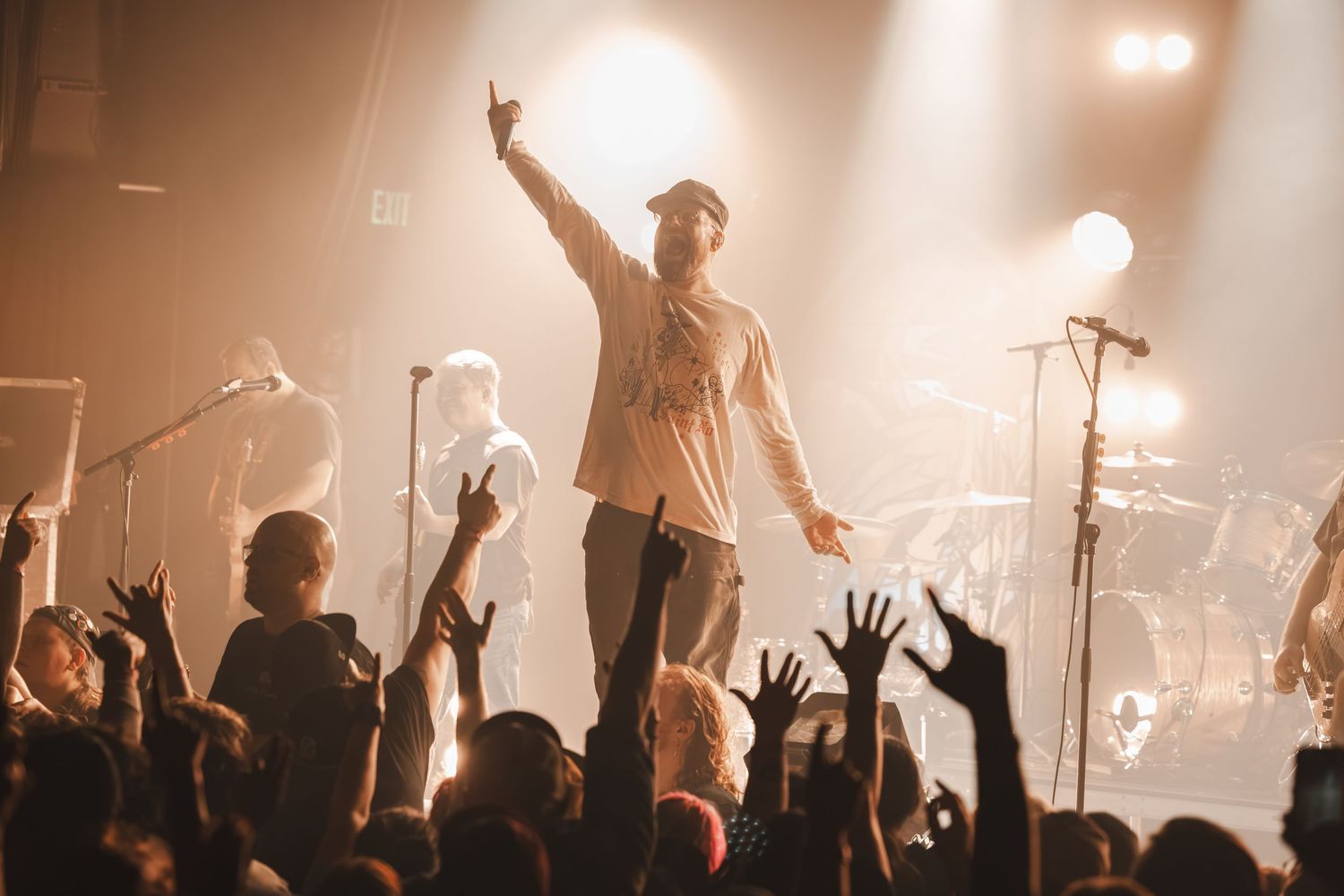 Energetic performer raises hand to crowd with band members visible on warmly lit concert stage.