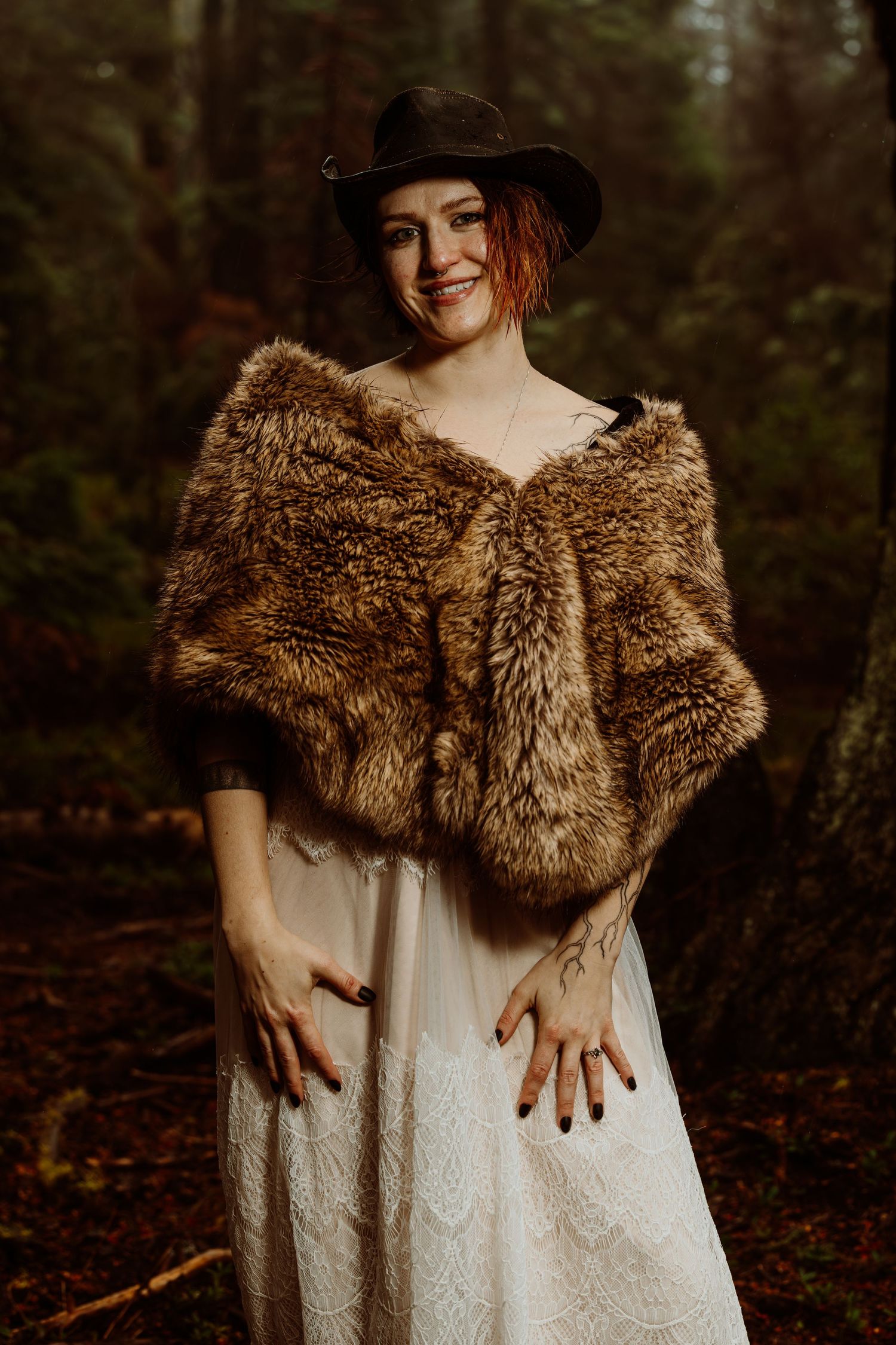 Smiling bride in white lace gown and brown fur wrap wearing black hat, standing in dark forest.