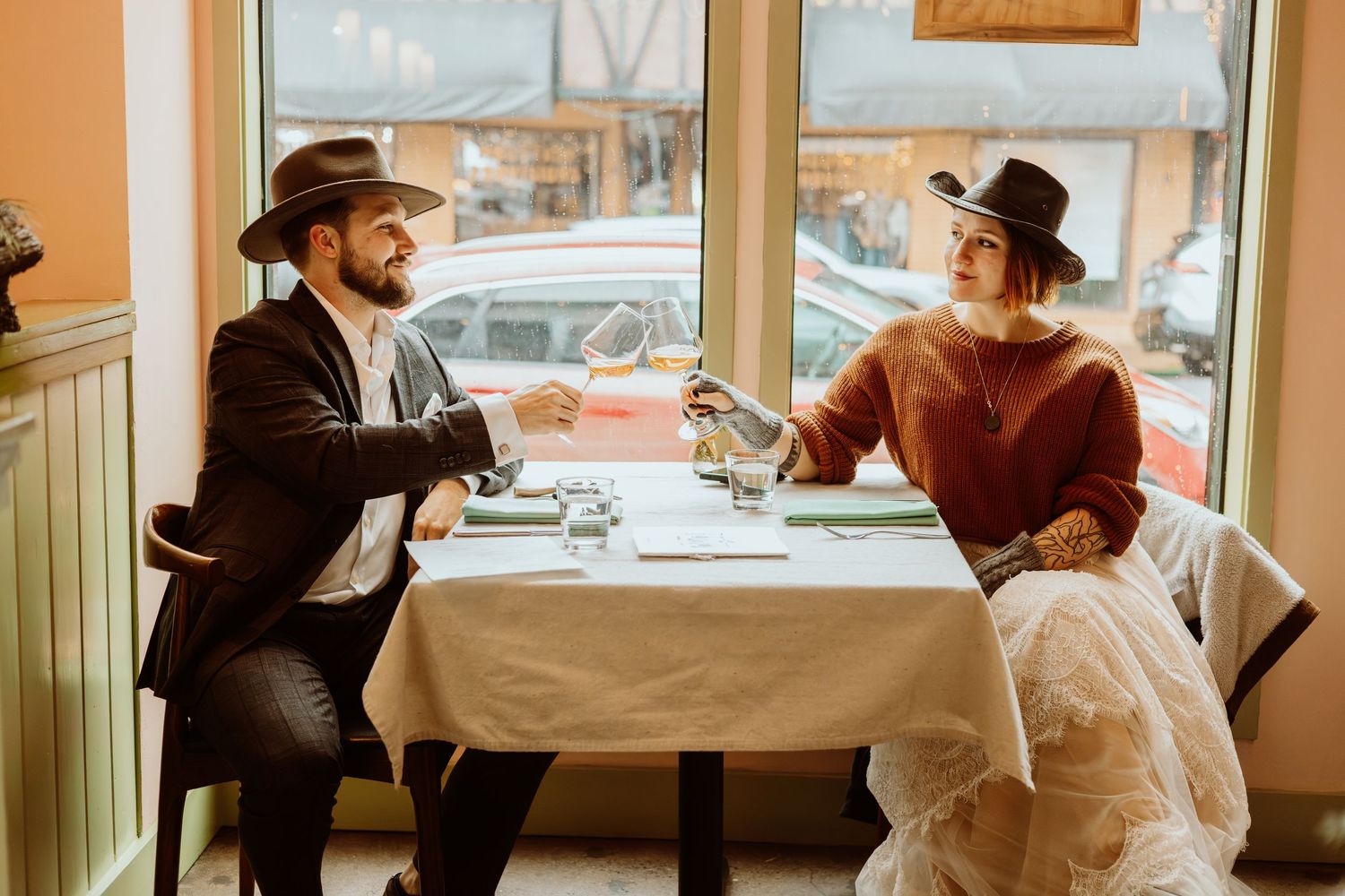 Newlywed couple in cowboy hats toasts with champagne at a restaurant table in casual western-inspired wedding attire.