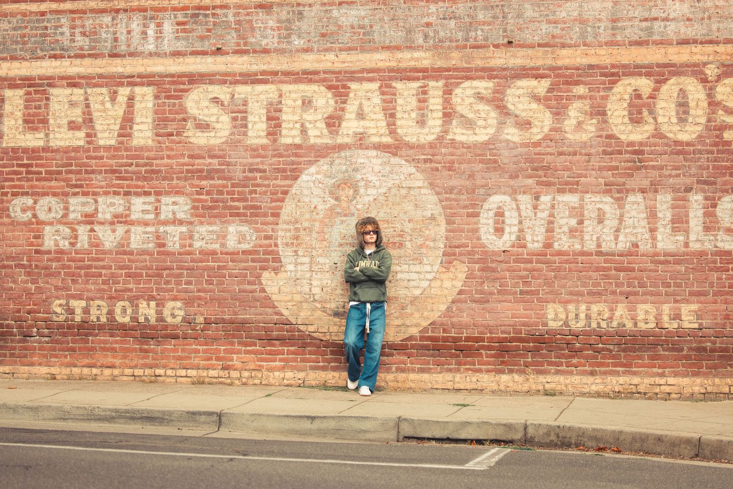 Teen in fur hat stands before iconic Levi Strauss and Co copper riveted overalls painted brick wall.