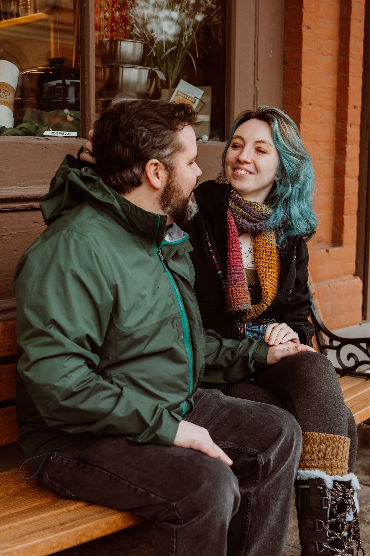 Laughing couple sitting together on a bench outside a shop, woman with teal hair and colorful scarf.