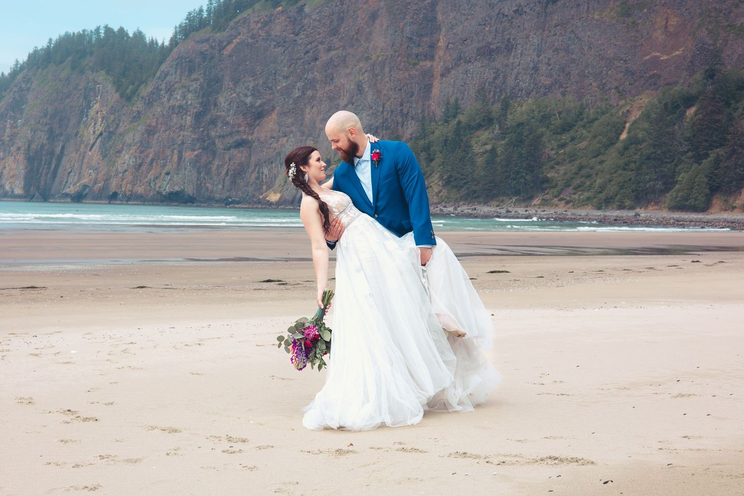 Groom dipping bride on a sandy beach with tall rocky cliffs and ocean waves behind them.