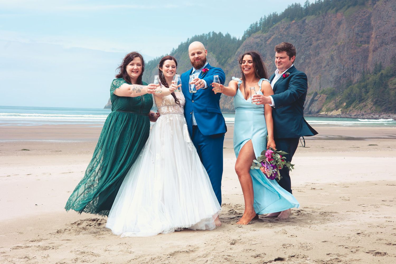 Wedding party of five celebrating with champagne on a sandy beach with coastal cliffs in background.
