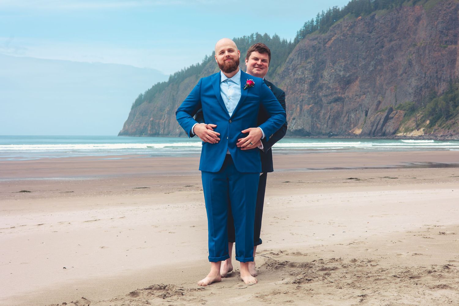 Two groomsmen in matching blue suits posing playfully barefoot on a sandy beach.